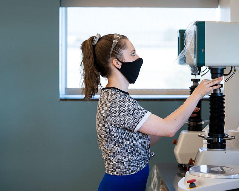 Girl in a lab with a mask on working on a piece of machinery.